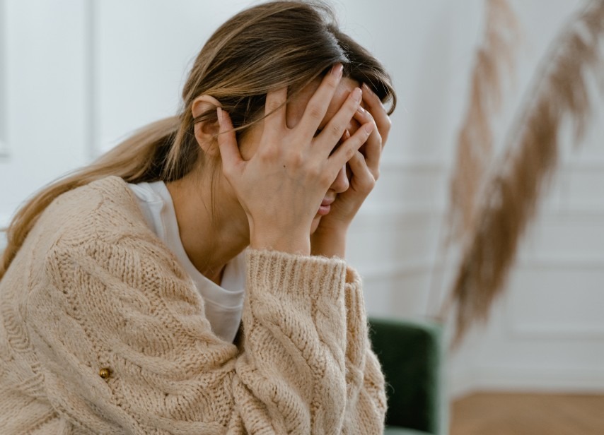 Woman wearing a beige cable-knit sweater, sitting indoors with her hand to her head