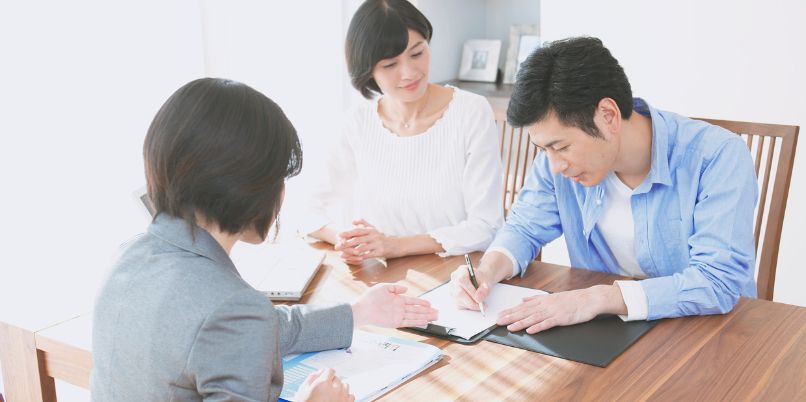 Three people sitting at a table, engaged in discussion while one person takes notes on a clipboard in a bright, welcoming setting