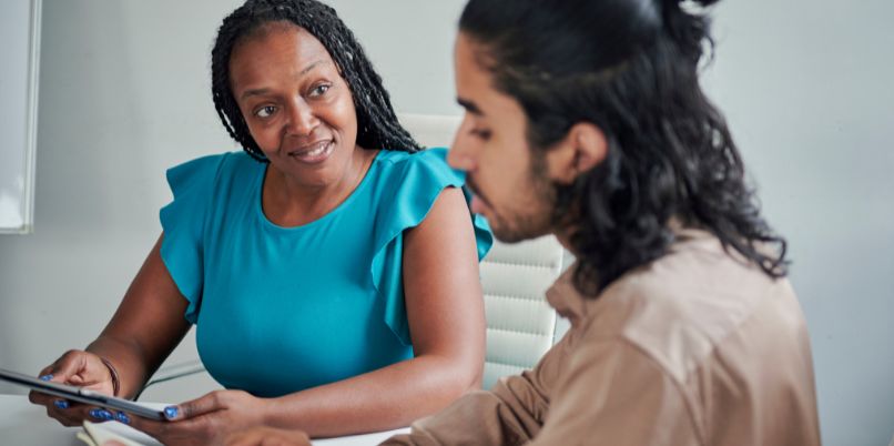 Two people sitting at a table having a discussion, one holding a tablet and the other listening attentively, with notebooks visible.