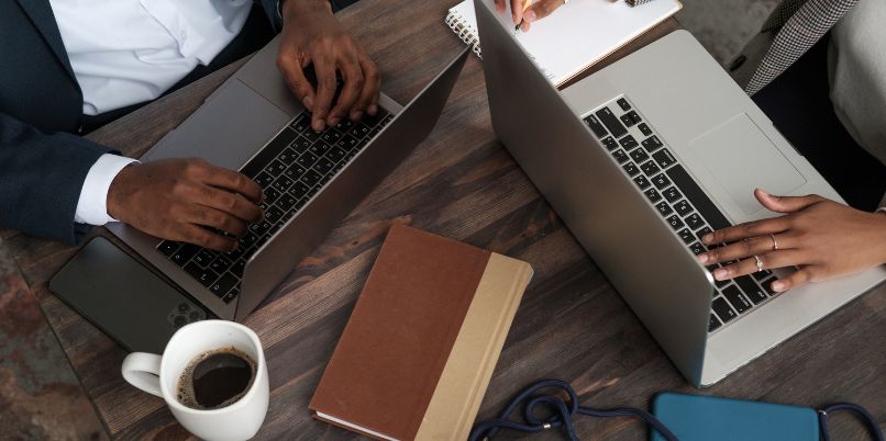 Two people working at a wooden table, each using a laptop, with notebooks, a cup of coffee, and a stethoscope visible