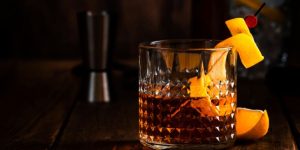 Glass of whiskey garnished with orange peel and a cherry, set on a dark wooden surface