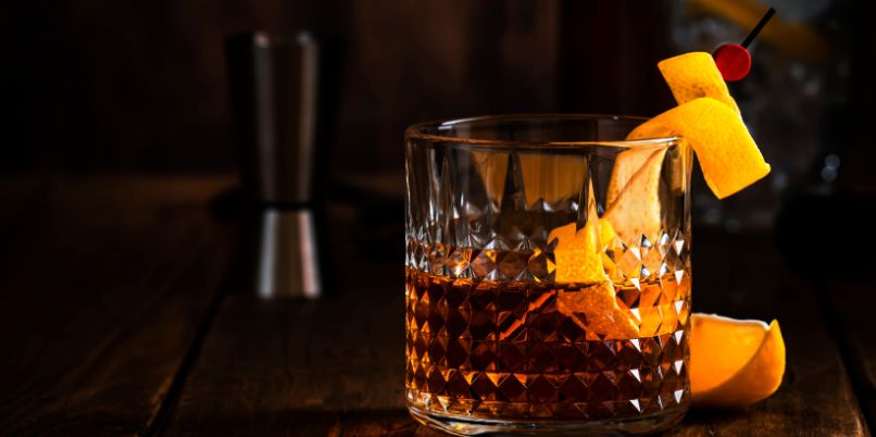 Glass of whiskey garnished with orange peel and a cherry, set on a dark wooden surface