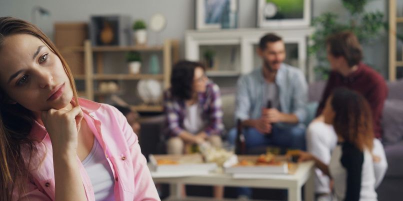 Young woman sitting apart and appearing thoughtful while a group of people socializes in the background of a cozy living room
