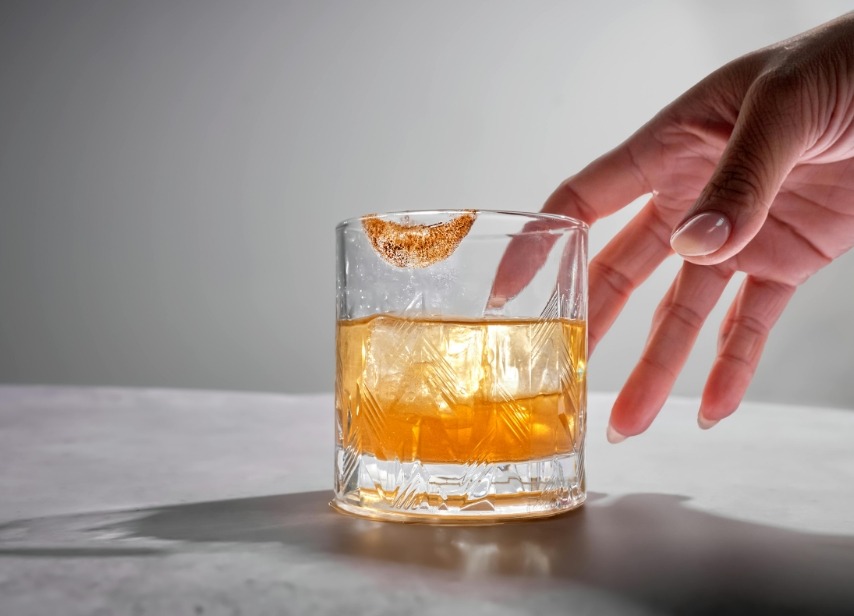 A hand reaching for a glass of whiskey on the rocks with a lipstick stain on the rim