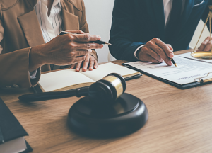 Two people in professional attire discussing legal documents at a desk, with a judge's gavel and legal scales visible