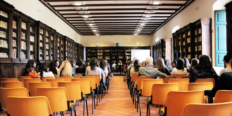 Audience seated in rows of orange chairs in a large library or lecture hall, attending a presentation at the front of the room