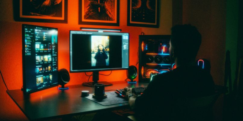 Person sitting at a computer desk illuminated by colorful LED lights, working on a multi-monitor setup in a dark room