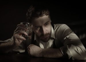 Man in a white dress shirt sitting at a table, holding a glass of whiskey in a dimly lit setting