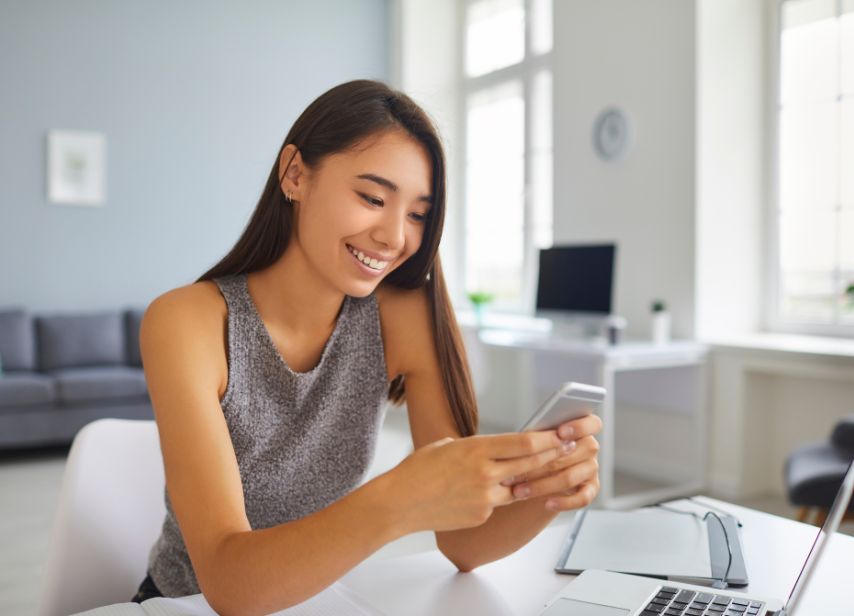 Woman sitting at a desk in a modern home office, holding a smartphone, with a laptop and glasses nearby