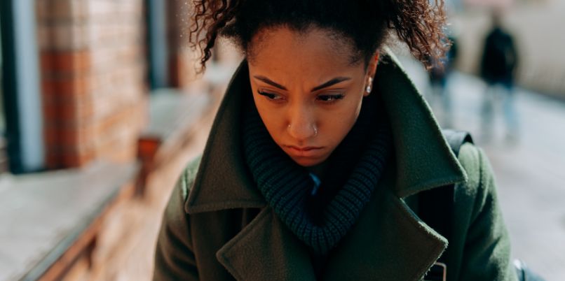 Person wearing a dark green coat and scarf walking outdoors, with focus on upper body and blurred city background