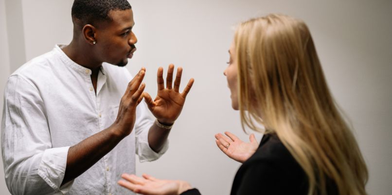 Two people in conversation, one gesturing with hands raised and the other listening, both standing indoors against a plain background.