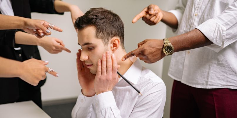 Stressed man in a white shirt sitting at a desk, surrounded by multiple hands pointing fingers at him in accusation.A man in a white shirt sits with his head in his hand while several people around him point fingers at him accusingly.