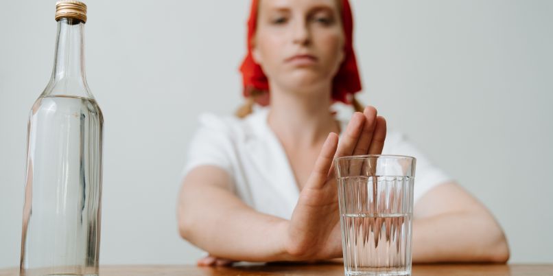 Person wearing a red headscarf and white shirt sitting at a table, making a stop gesture toward a glass and bottle of alcohol